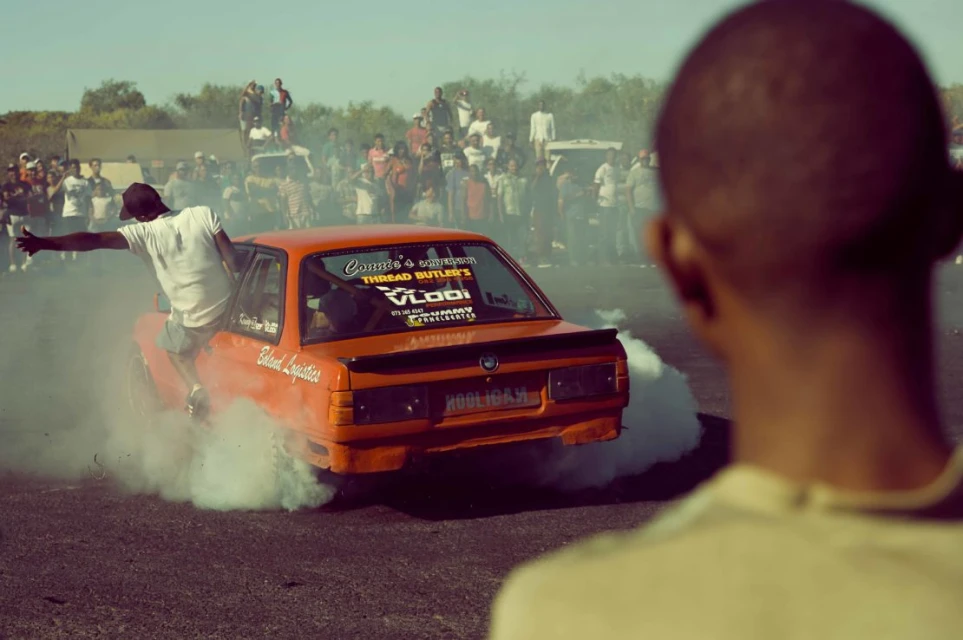 Orange sedan drifting on asphalt with a man hanging off the side door, surrounded by white smoke, and a crowd in the background.