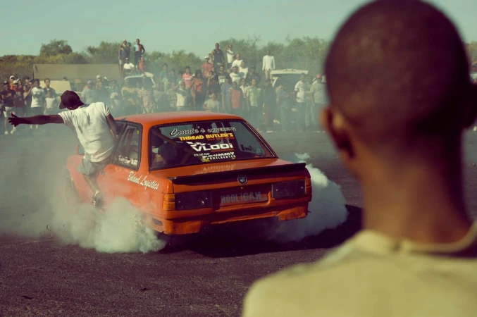 Orange sedan drifting on asphalt with a man hanging off the side door, surrounded by white smoke, and a crowd in the background.