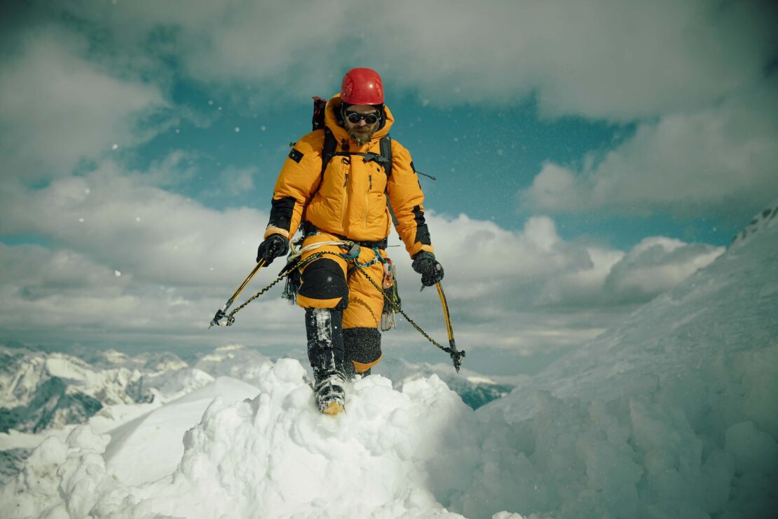 Mountain climber in yellow jacket ascends snowy peak with ice axes against a cloudy sky in a medium shot, demonstrating outdoor adventure.