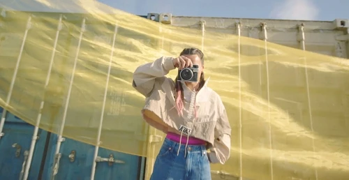 Adult woman in crop top and jeans, holds a camera and takes a picture outdoors in front of a yellow-draped building on a sunny day.
