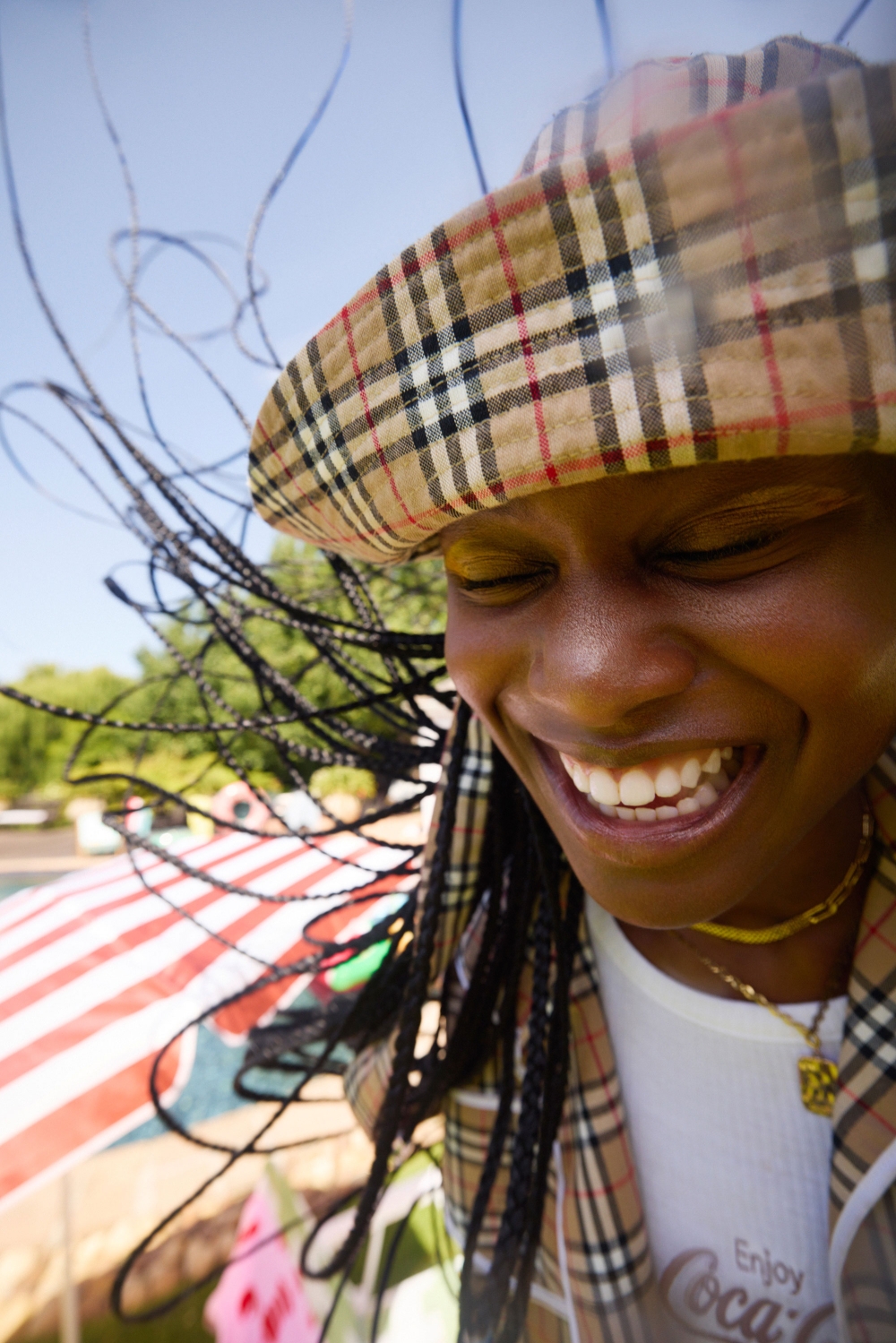 Close-up of a Black woman smiling with a plaid hat, braids, and a Coca-Cola t-shirt, outside on a sunny day with trees visible.
