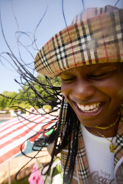 Close-up of a Black woman smiling with a plaid hat, braids, and a Coca-Cola t-shirt, outside on a sunny day with trees visible.