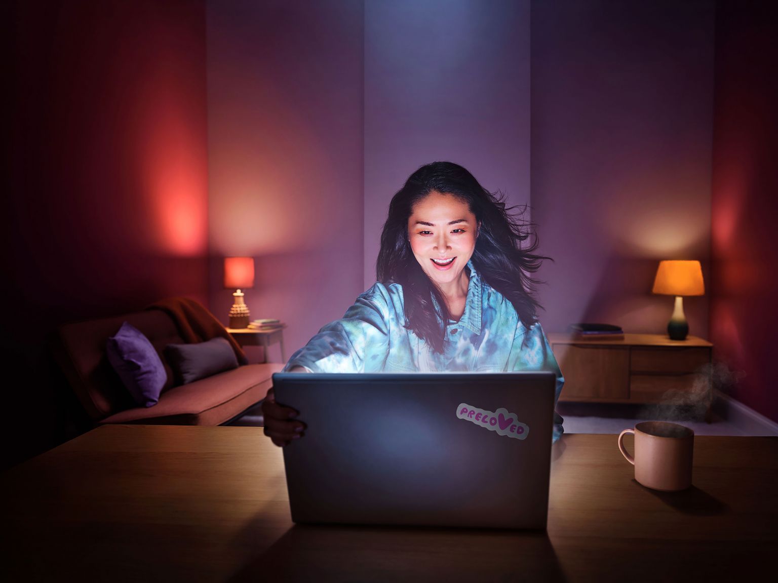 A smiling woman sits at a table in a room, looking at a laptop screen, with a cup of coffee and a warm, inviting atmosphere.