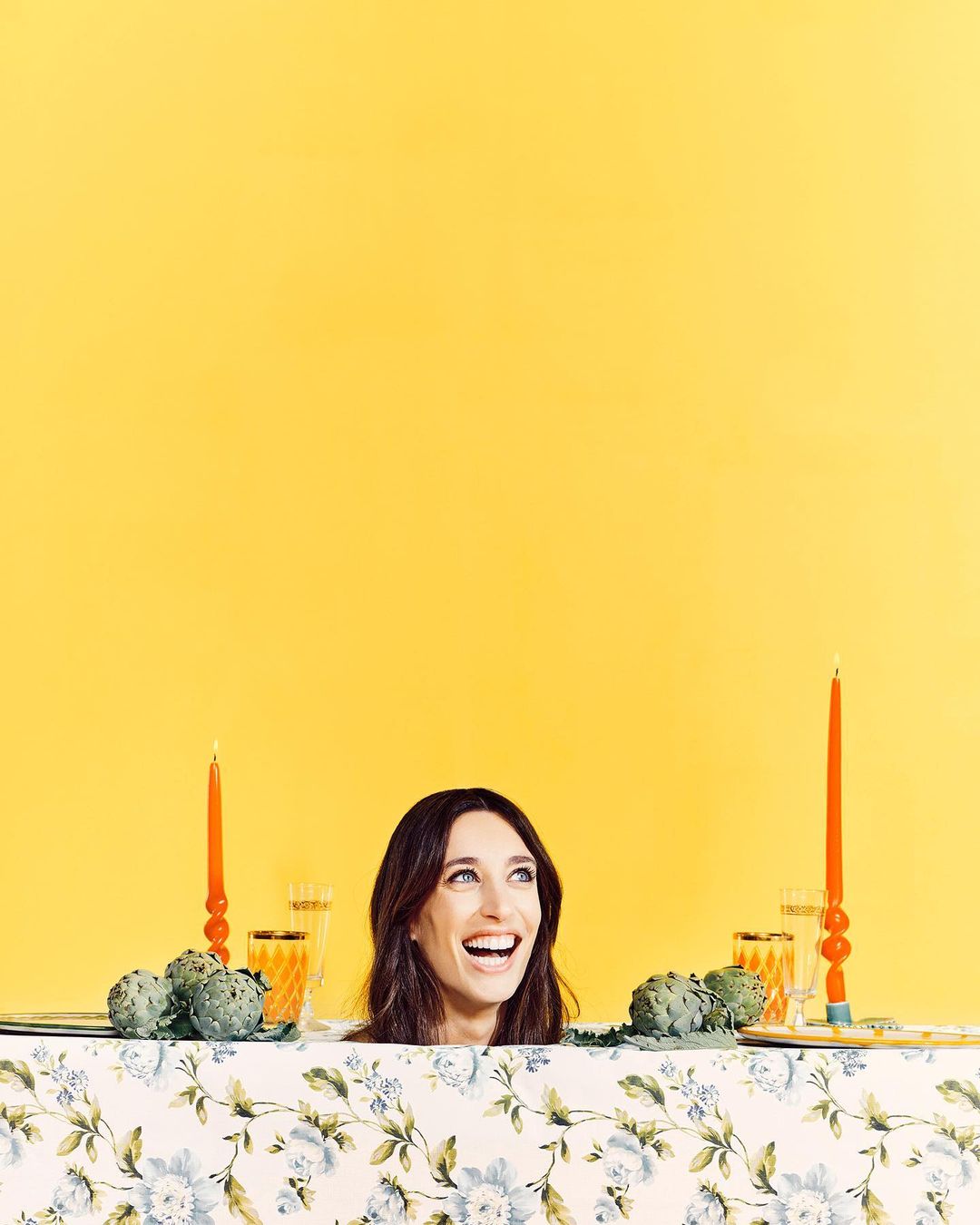 Portrait of smiling adult woman at a table decorated with artichokes, candles, and glassware against a vibrant yellow backdrop.