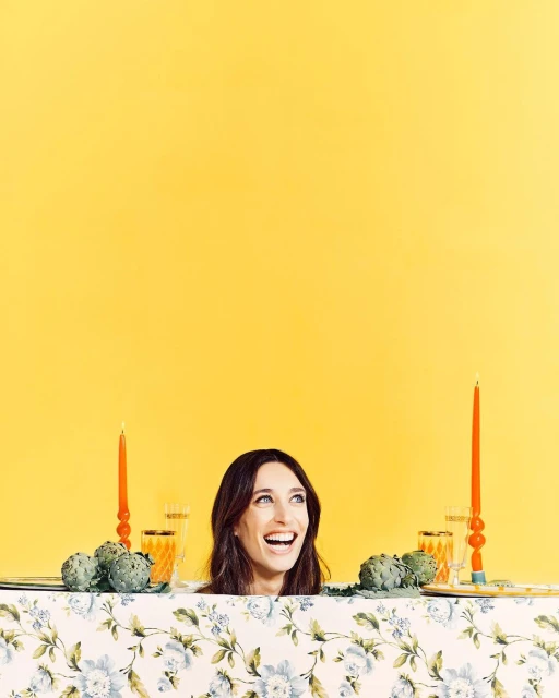 Portrait of smiling adult woman at a table decorated with artichokes, candles, and glassware against a vibrant yellow backdrop.
