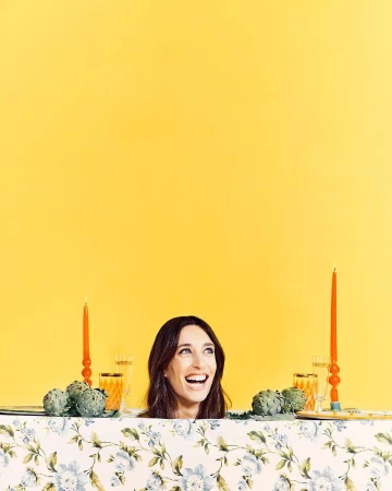 Portrait of smiling adult woman at a table decorated with artichokes, candles, and glassware against a vibrant yellow backdrop.