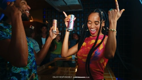 Two young adults dancing in a bar, holding canned drinks, with other people in the background under dim, party-like lighting conditions.