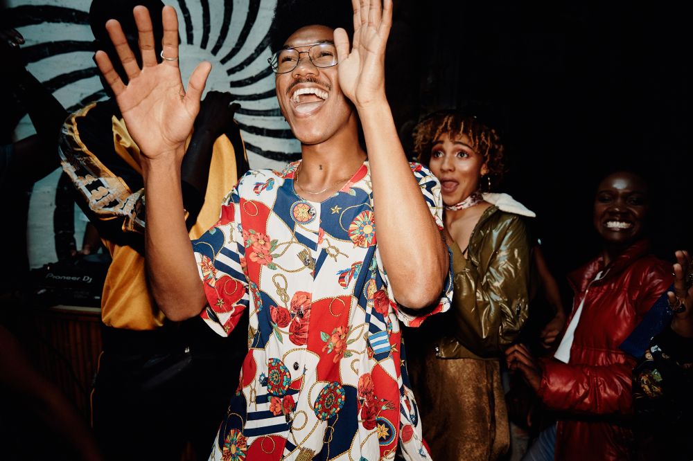 A lively indoor event featuring a group of stylishly dressed people, with one man in a patterned shirt raising his hands and smiling.