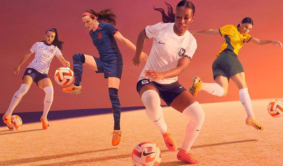 Four female soccer players in uniform skillfully juggle soccer balls, demonstrating their athleticism on a tan surface against an orange backdrop.