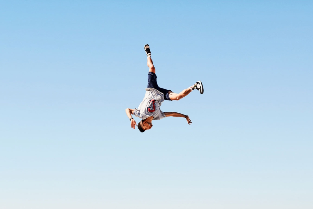 A man in mid-air performing an acrobatic stunt against a clear blue sky backdrop, showcasing fitness and outdoor activity.