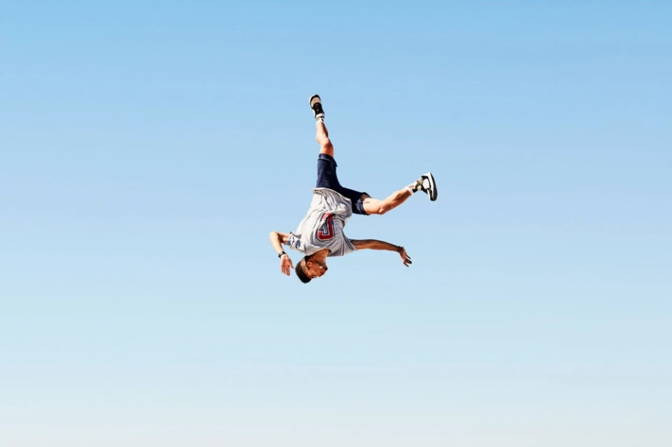 A man in mid-air performing an acrobatic stunt against a clear blue sky backdrop, showcasing fitness and outdoor activity.