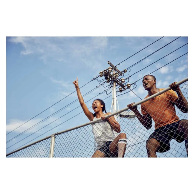 Two cheering athletes, an African American woman and man, are holding onto a fence during daytime with blue sky and power lines visible.