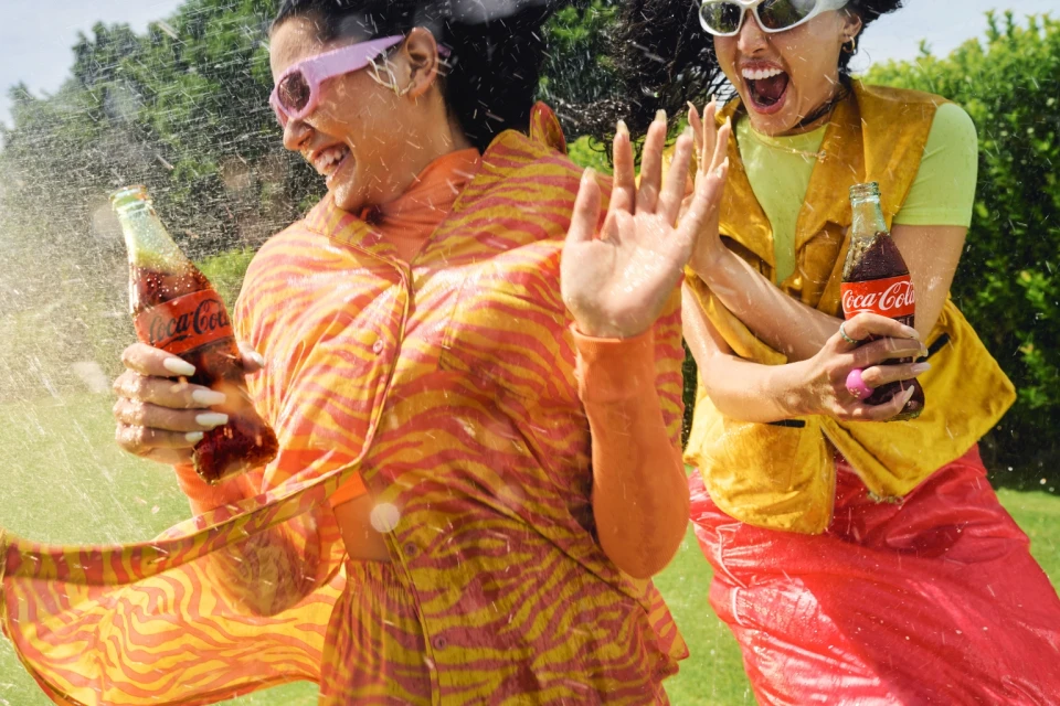 Two adult women in colorful outfits playfully hold Coca-Cola bottles, splashing water in an outdoor setting with vibrant green background.