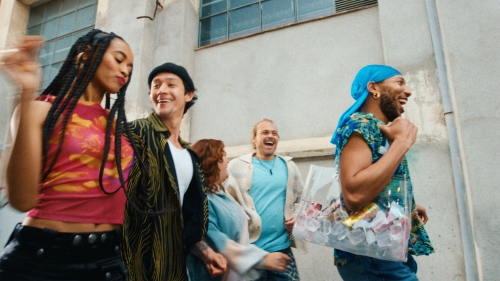 A group of five diverse friends laugh and walk together outdoors in an urban setting with bright daylight and a clear plastic tote bag.