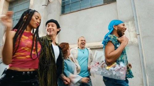 A group of five diverse friends laugh and walk together outdoors in an urban setting with bright daylight and a clear plastic tote bag.