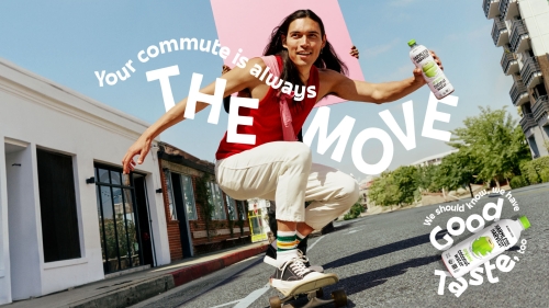 Adult male rides skateboard on city street while holding bottled beverage and pink board in outdoor shot under blue sky.