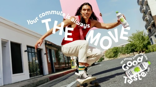 Adult male rides skateboard on city street while holding bottled beverage and pink board in outdoor shot under blue sky. 