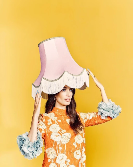 Woman in an orange floral dress balances a pink lampshade on her head against a yellow background in a studio setting, in a medium shot.
