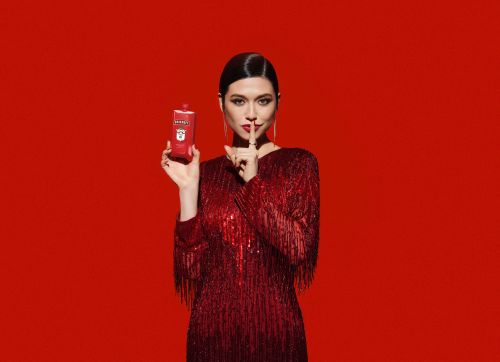 Elegant woman in red dress poses with a carton of beverage against a vibrant red backdrop, making a 'shh' gesture.