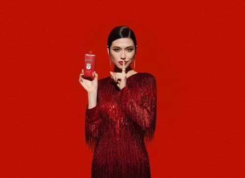 Elegant woman in red dress poses with a carton of beverage against a vibrant red backdrop, making a 'shh' gesture.