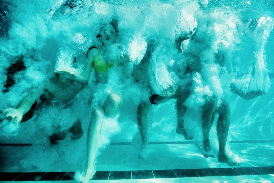 Underwater view of group swimming in a pool, bodies surrounded by bubbles with sunlight filtering through the clear turquoise water.