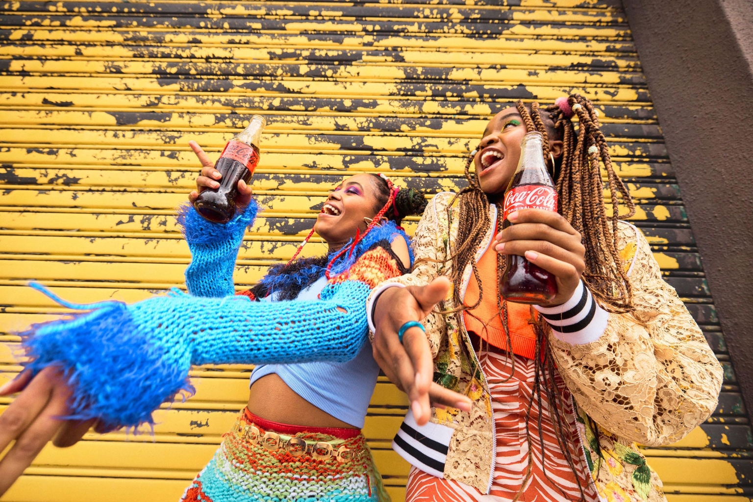 Two women smile and hold Coca-Cola bottles in front of a textured yellow wall, adorned in colorful, patterned clothing.