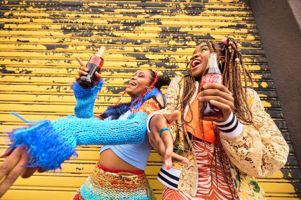 Two women smile and hold Coca-Cola bottles in front of a textured yellow wall, adorned in colorful, patterned clothing.