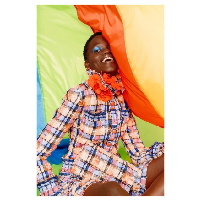 A smiling Black woman in a patterned tweed jacket with a bright orange flower collar, poses against a colorful draped background.