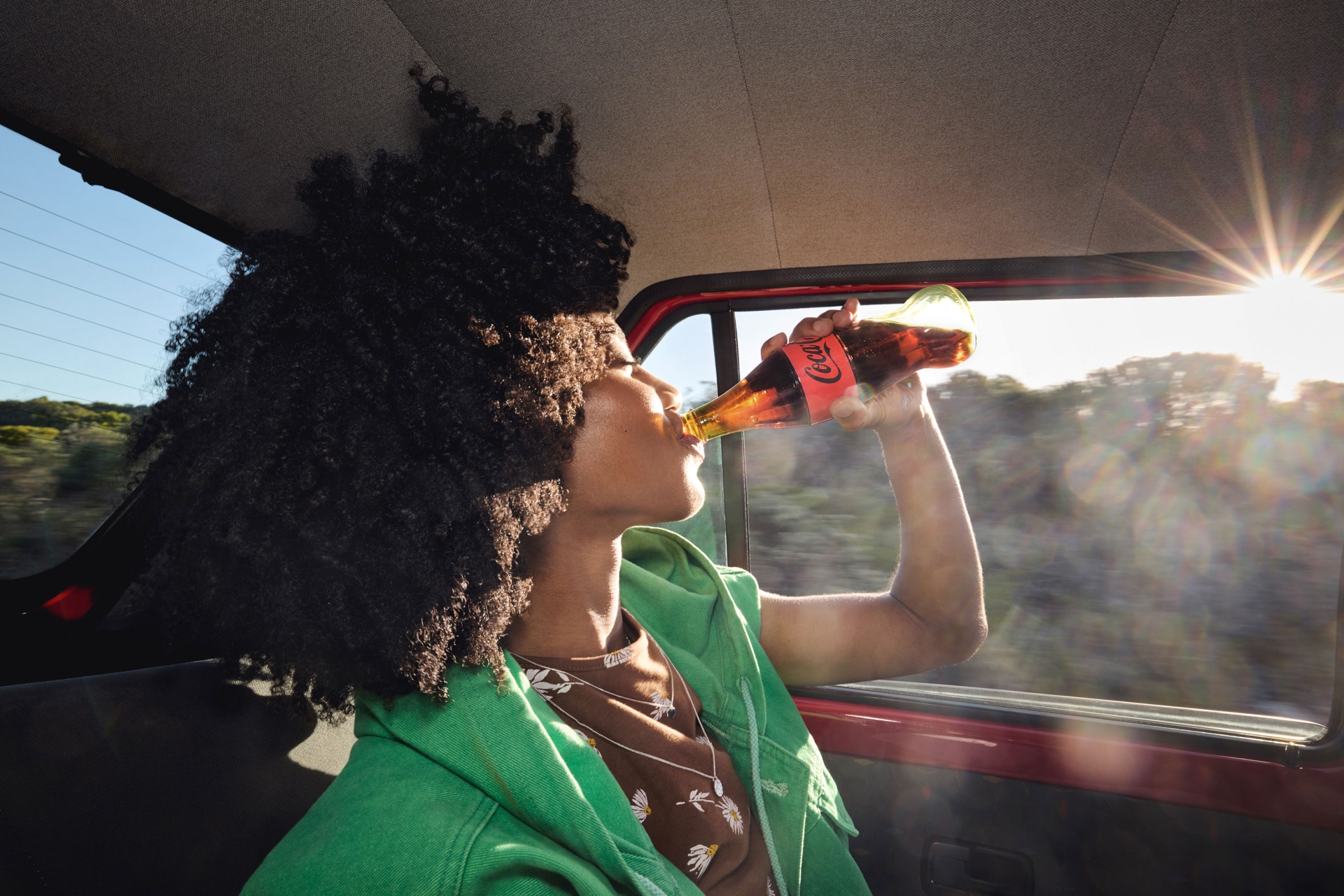 A young adult with curly hair drinks Coca-Cola in a car, wearing a green jacket, with sunlight creating a lens flare effect.