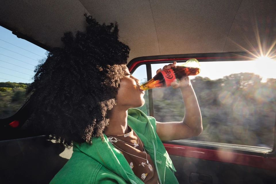 A young adult with curly hair drinks Coca-Cola in a car, wearing a green jacket, with sunlight creating a lens flare effect.