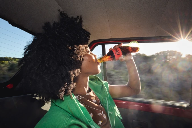 A young adult with curly hair drinks Coca-Cola in a car, wearing a green jacket, with sunlight creating a lens flare effect.