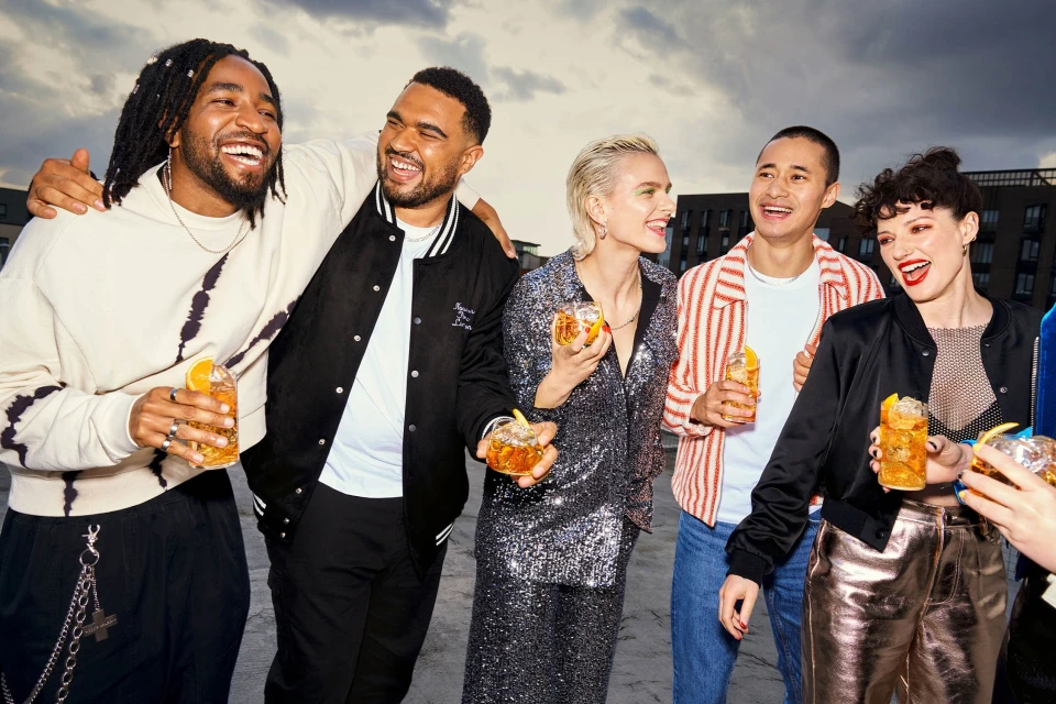 Five young adults smiling, dressed in jackets and shiny clothing, hold drinks garnished with orange slices on a rooftop at dusk.