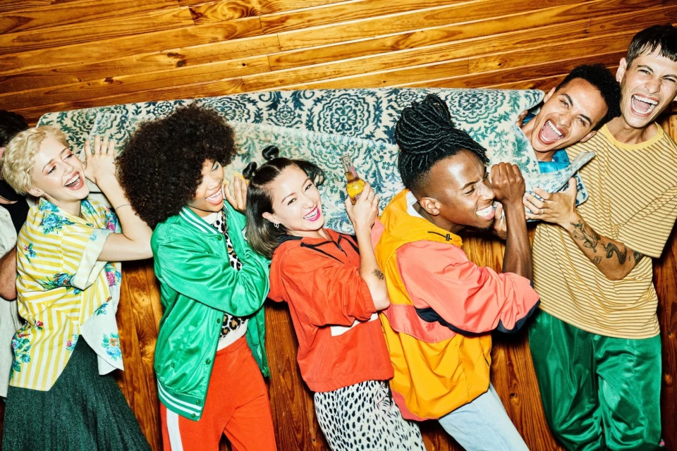 Group of six young adult friends laughing and holding a person on a couch against a wood paneled wall in an indoor setting.