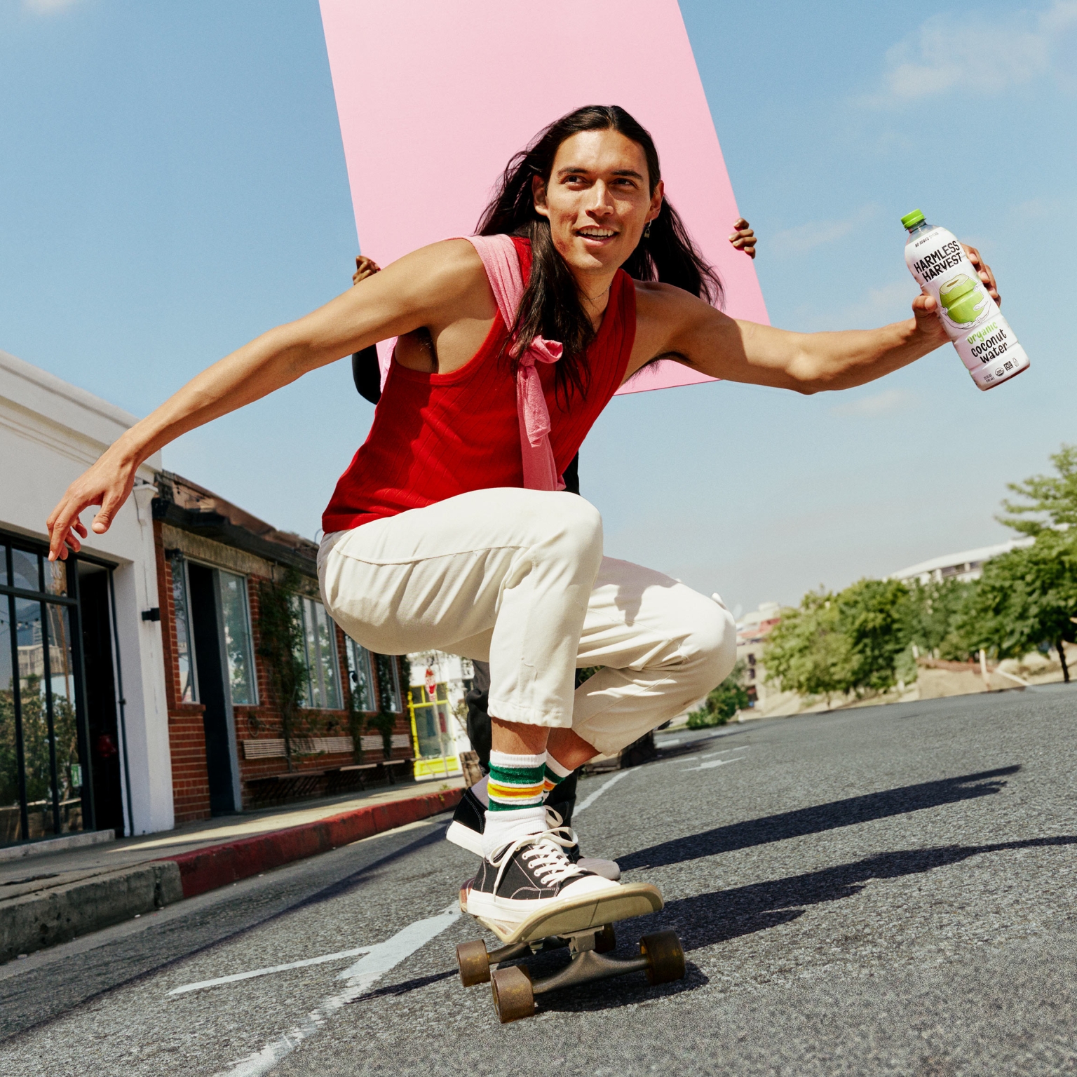 Young man with long dark hair balances on a skateboard outdoors, holding a bottle of coconut water and a pink poster in a bright urban scene.