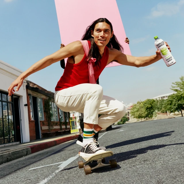 Young man with long dark hair balances on a skateboard outdoors, holding a bottle of coconut water and a pink poster in a bright urban scene.