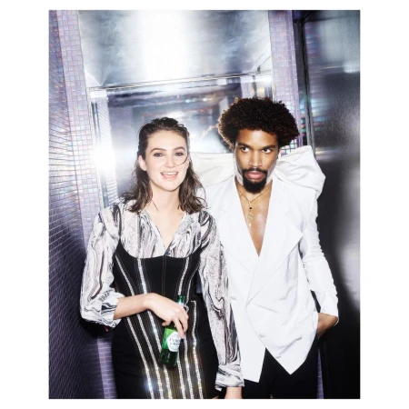 Portrait of a smiling woman holding a green bottle and a Black man in a white blazer posing indoors by Tom Van Schelven