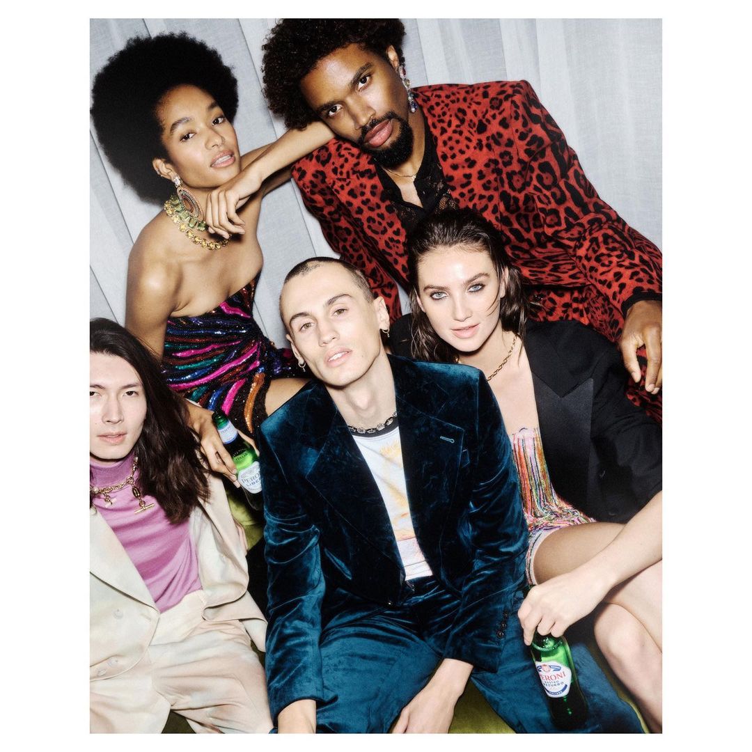 Group of five diverse young adults in stylish party attire pose with bottles. Includes African-American and Asian individuals in indoor setting.