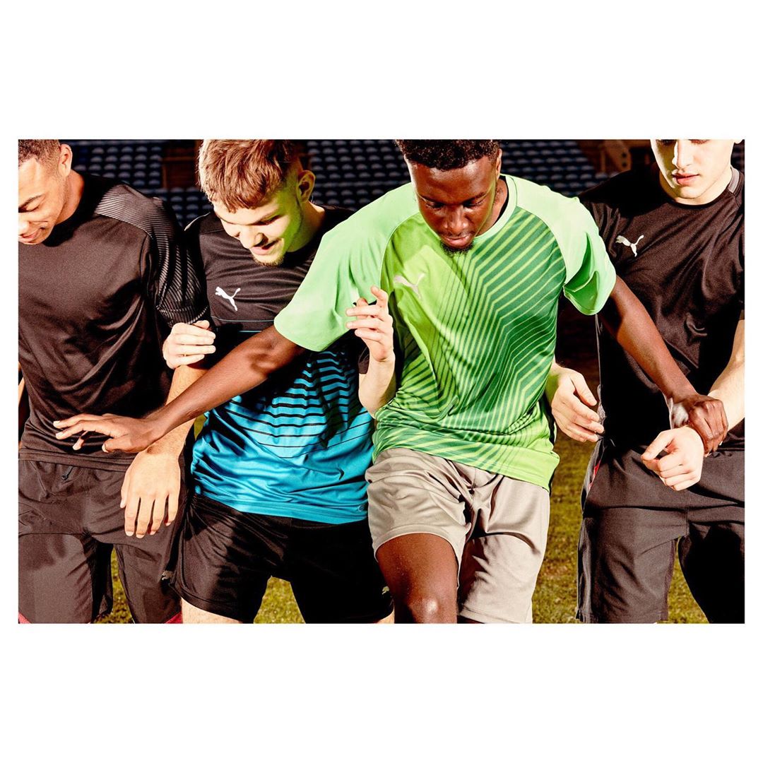 Four male athletes stand together outdoors, wearing athletic shirts and shorts, with grass visible in the background and natural light.