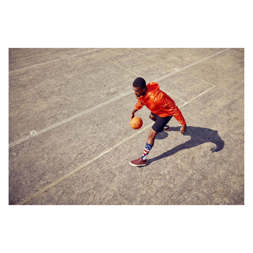 An African-American adult male bounces a basketball on a concrete court wearing a bright orange jacket and striped socks from an eye-level angle.