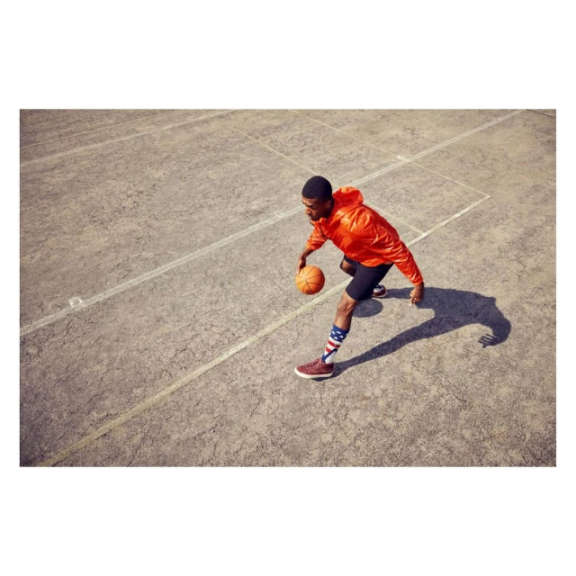An African-American adult male bounces a basketball on a concrete court wearing a bright orange jacket and striped socks from an eye-level angle.