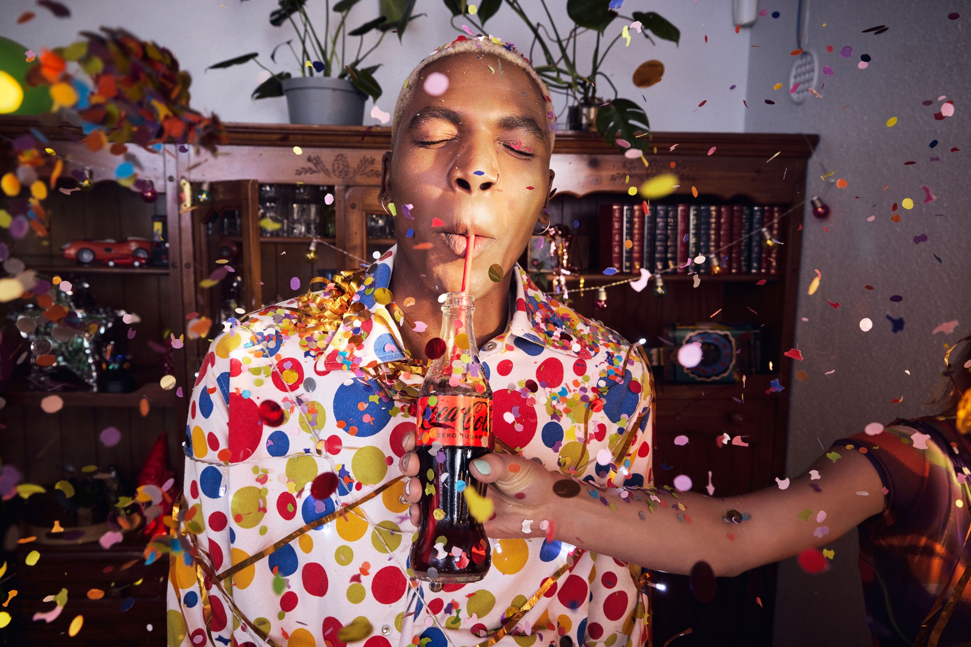 Person drinking Coca-Cola through a straw with confetti falling, wearing a polka dot shirt in an indoor environment.