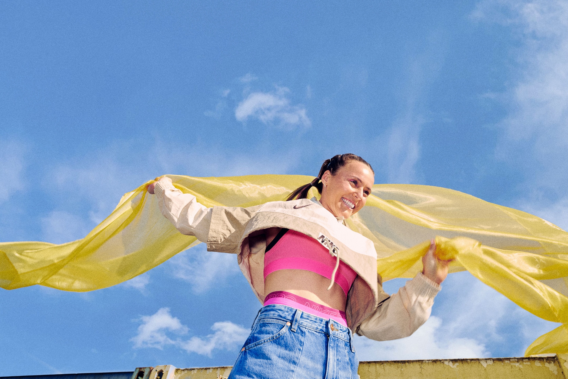 Smiling woman in pink sports bra and denim skirt holds a yellow scarf against a blue sky, wearing a Nike jacket with a logo visible.