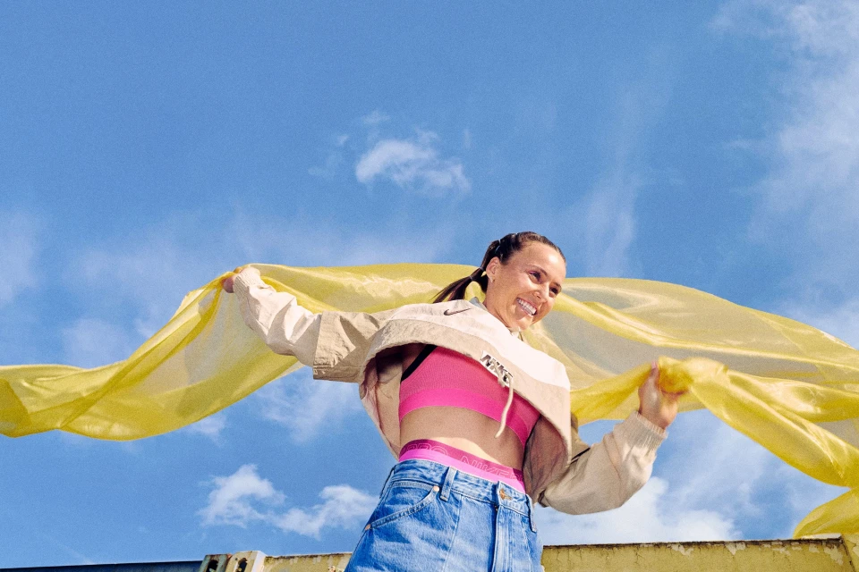 Smiling woman in pink sports bra and denim skirt holds a yellow scarf against a blue sky, wearing a Nike jacket with a logo visible.