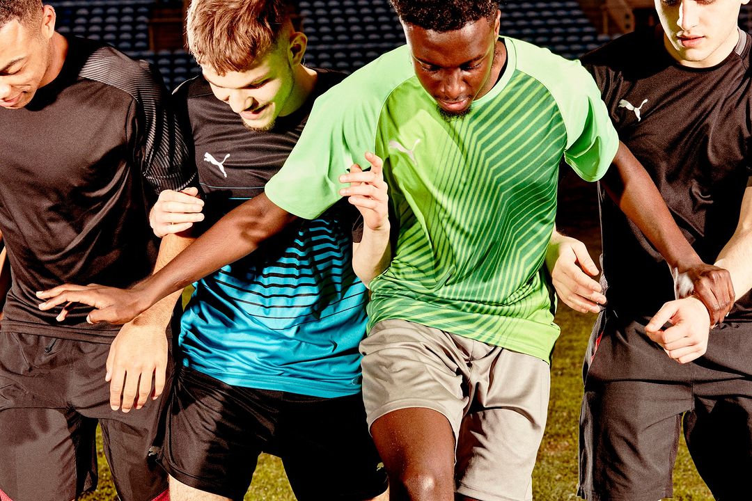A group of four young adult men of diverse ethnicities stand outdoors on a sports field wearing athletic shirts. They are standing close to one another.