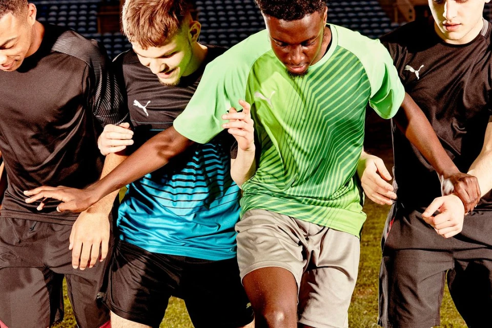 A group of four young adult men of diverse ethnicities stand outdoors on a sports field wearing athletic shirts. They are standing close to one another.
