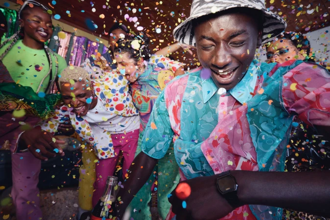 Group of smiling young adults celebrating a party with colorful confetti, wearing patterned clothing in an indoor environment.