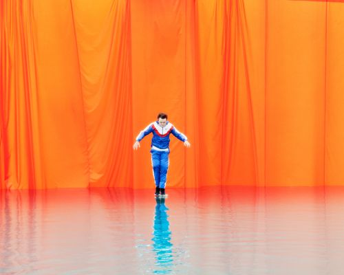 An adult male in a blue tracksuit stands in front of an orange backdrop, his reflection shimmering on a glossy surface below.