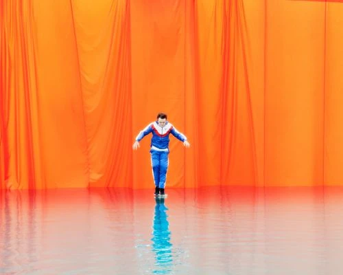 An adult male in a blue tracksuit stands in front of an orange backdrop, his reflection shimmering on a glossy surface below.