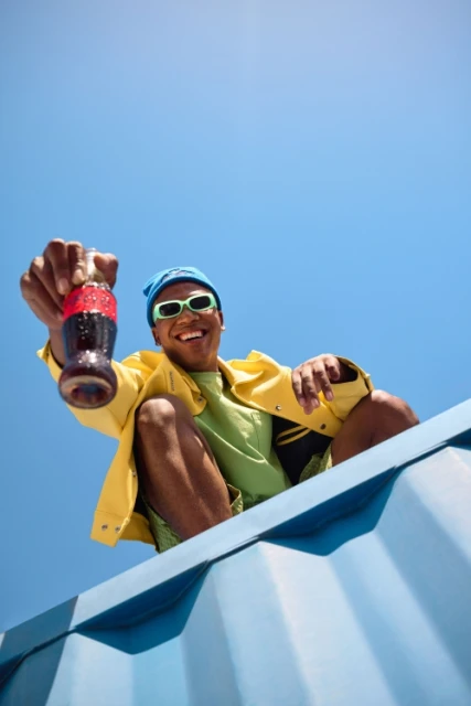 Young Black man smiles, offering a Coca-Cola bottle. He's wearing a yellow jacket, green shirt, blue bandana, and sunglasses against a blue sky.