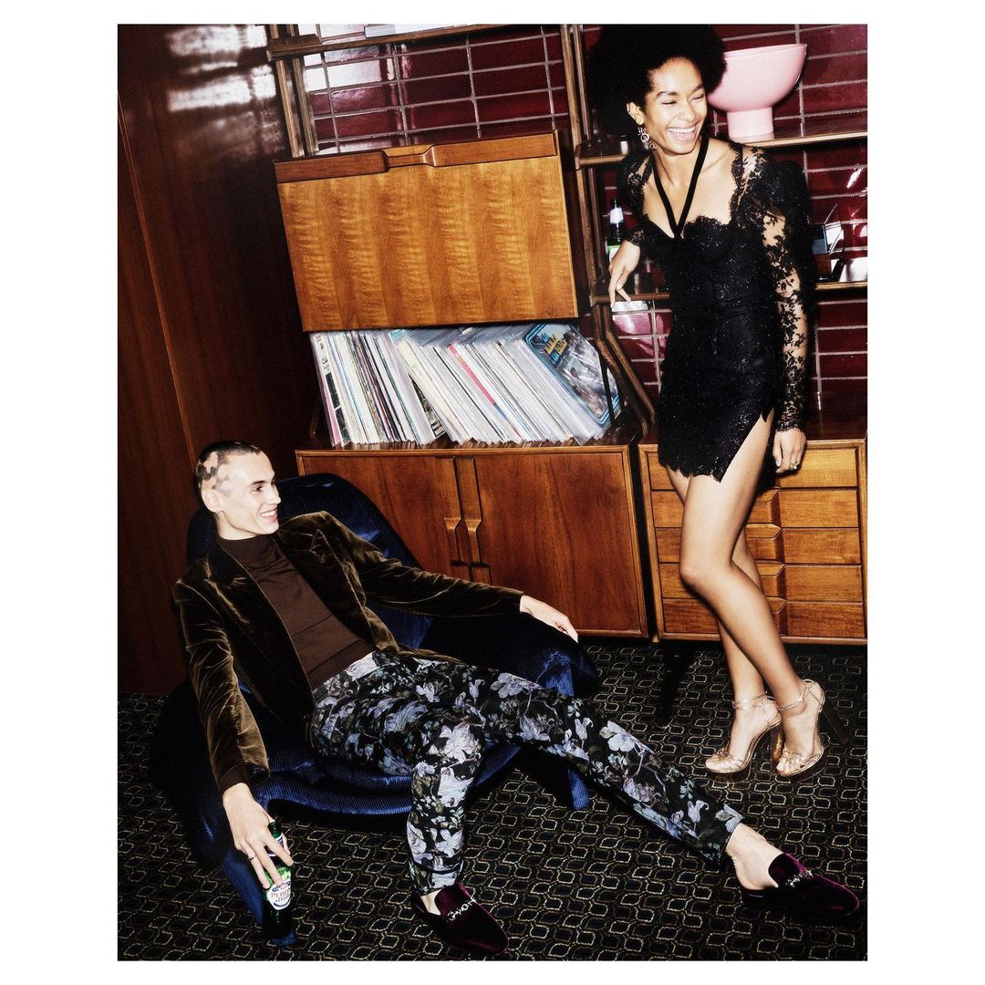 Stylish indoor shot of a woman in a short black dress and a man in a velvet blazer, with a mid-century cabinet and bar accessories.